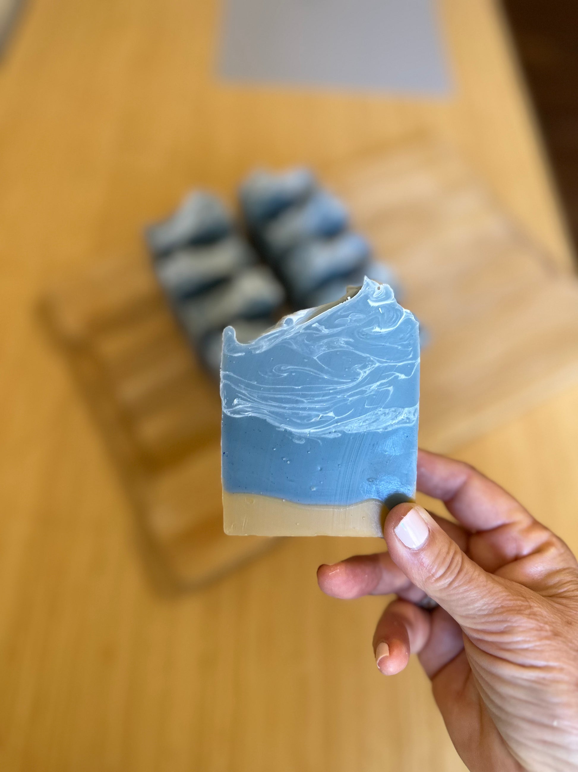 Hand holding a handmade artisan soap bar with blue and cream layers, featuring a textured top. Natural, vegan cold process soap with a coastal-inspired design. Blurred background with additional soap bars on a wooden board