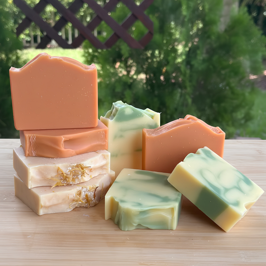 Five artisanal bars of soap in various colors and patterns, including solid orange, marbled green and cream, and layered beige, arranged on a wooden board with a blurred garden background.