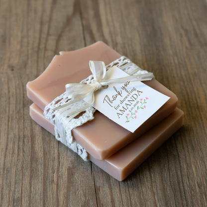 A rectangular bar of blush pink soap wrapped with a cream lace ribbon and tied with a matching ribbon bow, featuring a small white thank-you card that reads 'Thank you for choosing AMANDA' with a small floral design. The soap is placed on a textured beige linen background.