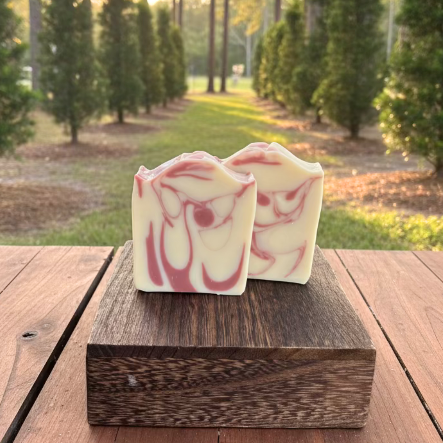 Two rectangular handmade soap bars with a cream and pink marbled swirl pattern, displayed on a dark wooden block on an outdoor wooden table.