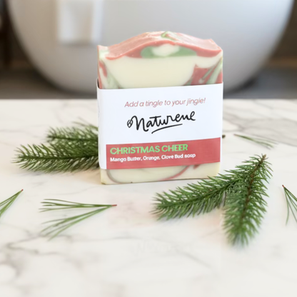 Bar of soap with a 'Naturene' label and Christmas cheer scent, surrounded by greenery on a light surface.